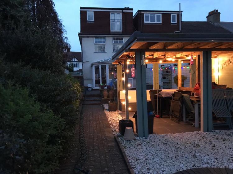 A picture of a back garden in the evening that includes a wooden pergola and outdoor garden lighting.